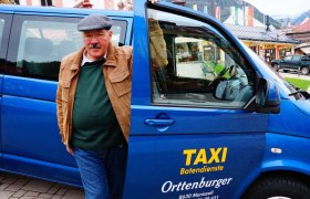 Taxi Orttenburger, © Ernst Miglbauer Ein Mann steht neben einem blauen Taxi mit der Aufschrift 'Taxi Botendienste Orttenburger'.