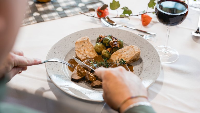 Regionale Köstlichkeit, © Niederösterreich Werbung/ David Schreiber Person schneidet Fleisch auf einem Teller mit Knödeln und Rosenkohl, daneben ein Glas Rotwein.