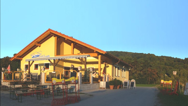 Florian's Teichstüberl, © Teichstüberl Ein gelbes Gebäude mit Terrasse und Biergartenmöbeln, umgeben von grünen Hügeln unter klarem Himmel.