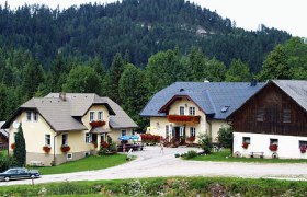 Drei Häuser in ländlicher Umgebung mit Wald im Hintergrund.