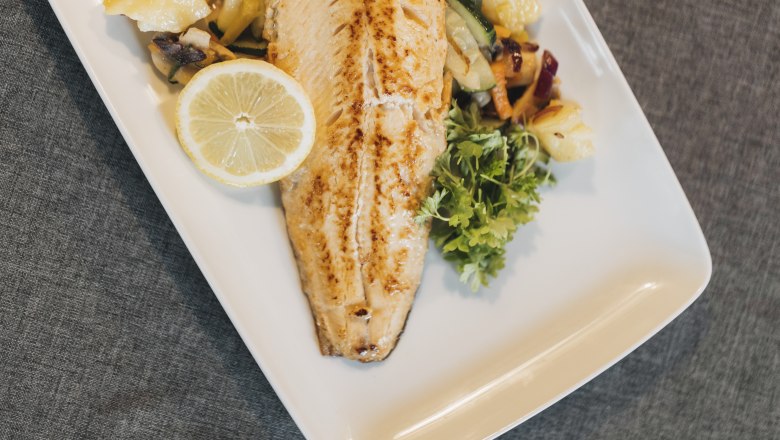 Saiblingsfilet mit Gemüse und Zitronenscheibe auf einem weißen Teller.