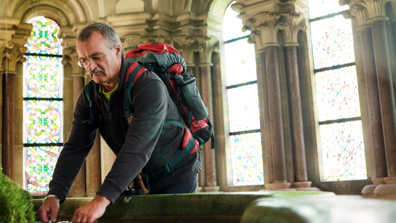 Ein Mann mit Rucksack beugt sich &uuml;ber ein Wasserbecken in einem historischen Geb&auml;ude mit bunten Glasfenstern.