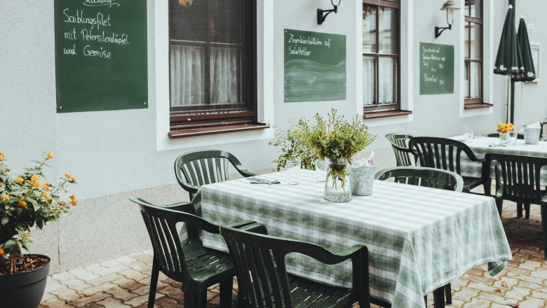 Ein Gastgarten mit grünen Stühlen und karierten Tischdecken vor einer Hauswand mit Kreidetafeln.