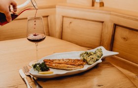 Ein Teller mit Zanderfilet und Kartoffeln in Knoblauchbutter, daneben ein Glas Rotwein auf einem Holztisch.