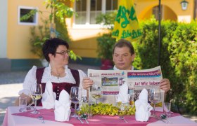 Ein Mann und eine Frau sitzen an einem Tisch im Freien, der mit Gläsern und Besteck gedeckt ist. Der Mann liest eine Zeitung.