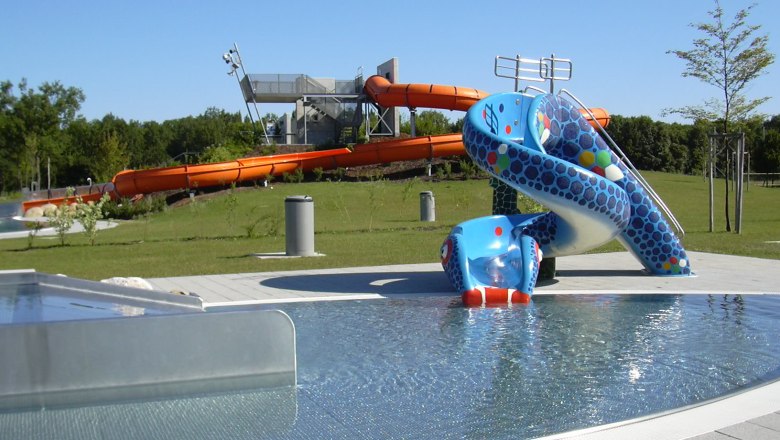 Blick auf einen Aquapark mit blauer Kinderrutsche und gro&szlig;er oranger Wasserrutsche im Hintergrund.