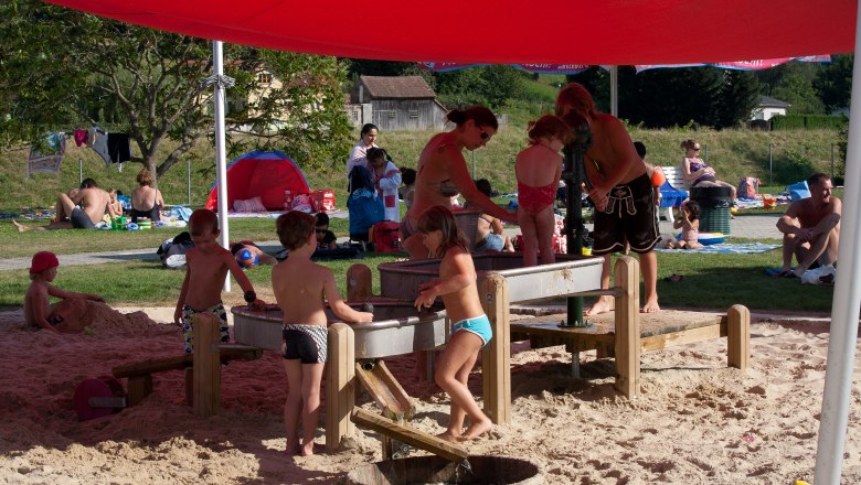 Kinder spielen im Sand unter einem roten Sonnensegel im Parkbad Wilhelmsburg.