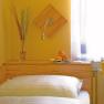 Gemütliches Zimmer mit gelben Wänden, Bett, Vase mit Gräsern und Obstschale auf Holzregal.