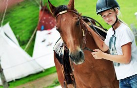 Ein M&auml;dchen mit Helm steht neben einem braunen Pferd auf einem Bauernhof.