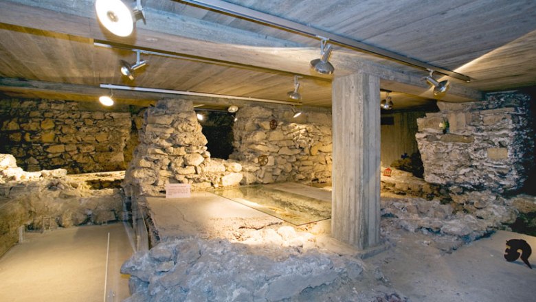 Unterirdische Ruinen mit Steinmauern und Betonstützen, beleuchtet durch Deckenstrahler.