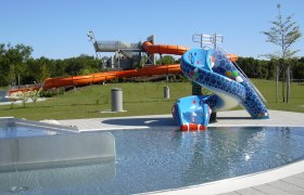 Blick auf einen Aquapark mit blauer Kinderrutsche und gro&szlig;er oranger Wasserrutsche im Hintergrund.
