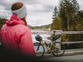 Ein Radfahrer genie&szlig;t die frische Luft und die atemberaubende Aussicht am Hubertussee. Umgeben von dichten W&auml;ldern und sanften H&uuml;geln, l&auml;dt die idyllische Landschaft zu einer entspannenden Pause ein. Die ruhige Wasseroberfl&auml;che spiegelt die Wolken und die Natur wider, w&auml;hrend die sanften Ger&auml;usche der Umgebung eine friedliche Atmosph&auml;re schaffen.