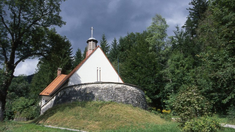 Kleine Kirche im Wald mit steinernem Sockel und rotem Dach.